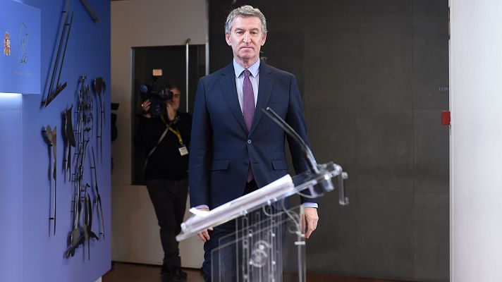 Feijóo, en traje azul oscuro, camisa blanca y corbata morada, camina hacia un atril, siendo grabado por un cámara.