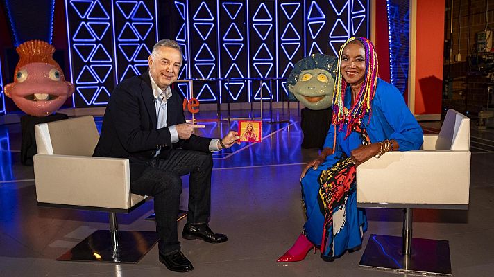 Entrevista en plató de televisión entre Cayetano Martínez de Irujo y Lucrecia.  Ella viste un vestido azul y él un traje oscuro; una marioneta verde está entre ellos.
