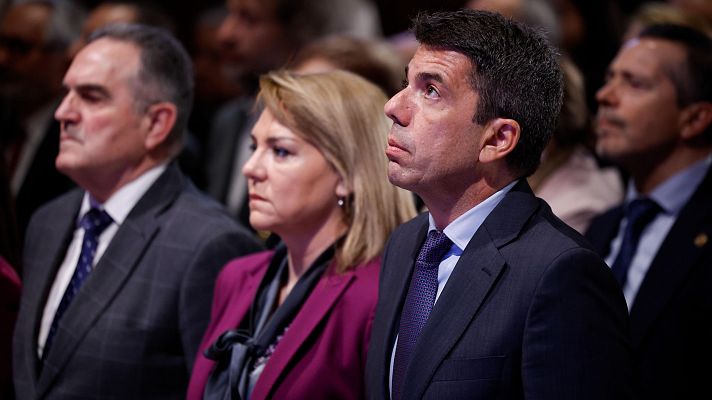 Grupo de personas en traje formal, posiblemente en un evento institucional. Un hombre con traje oscuro y corbata azul mira hacia arriba con atención.