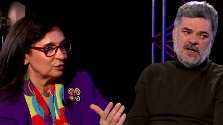 En un estudio oscuro, una mujer con gafas y chaqueta morada y un hombre con barba canosa participan en un programa llamado \"Voces Maestras\", relacionado con la dirección de programación y contenidos.