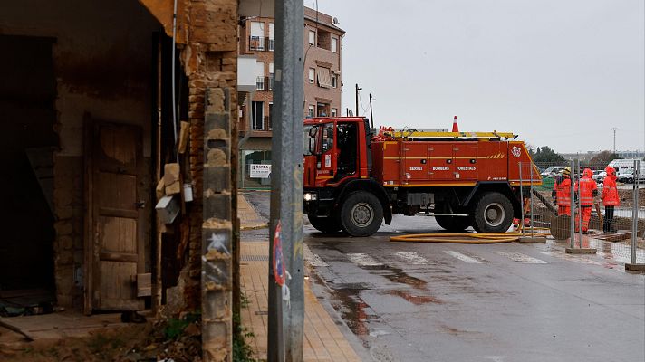 Camión de bomberos rojo en calle mojada, bomberos con trajes naranjas trabajan con una manguera.  Daños visibles en edificios cercanos.