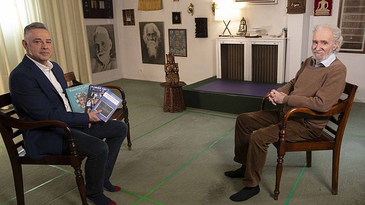 Dos hombres, uno en traje azul y otro en jersey marrón, sentados en sillas de madera en una habitación con decoración oriental.  Uno sostiene libros, uno de ellos con su nombre.