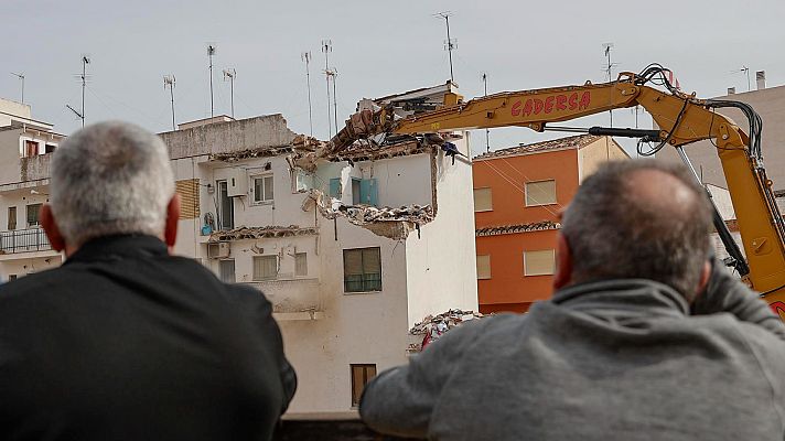Demolición de edificio blanco dañado con excavadora. Dos hombres observan la escena. Edificio naranja cercano sin daños.