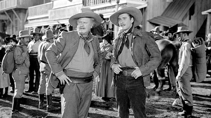 Imagen en blanco y negro de una escena de película del oeste con dos personajes principales, uno con traje a cuadros y otro con chaqueta de ante, rodeados de gente y edificios de madera.