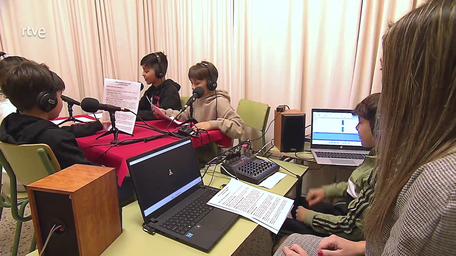 Niños leen noticias en una sesión de radio educativa, usando micrófonos y auriculares, con supervisión adulta y equipo de audio profesional.