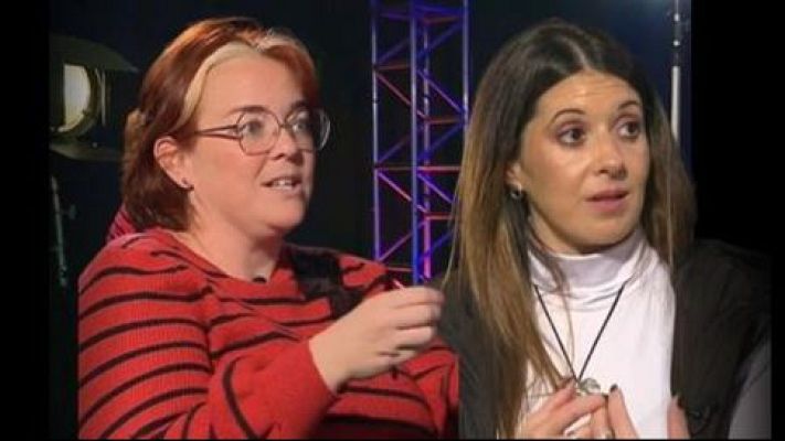 Entrevista o conversación entre dos mujeres con jerseys de rayas rojas y cuello alto blanco, respectivamente.  Fondo oscuro con iluminación profesional.