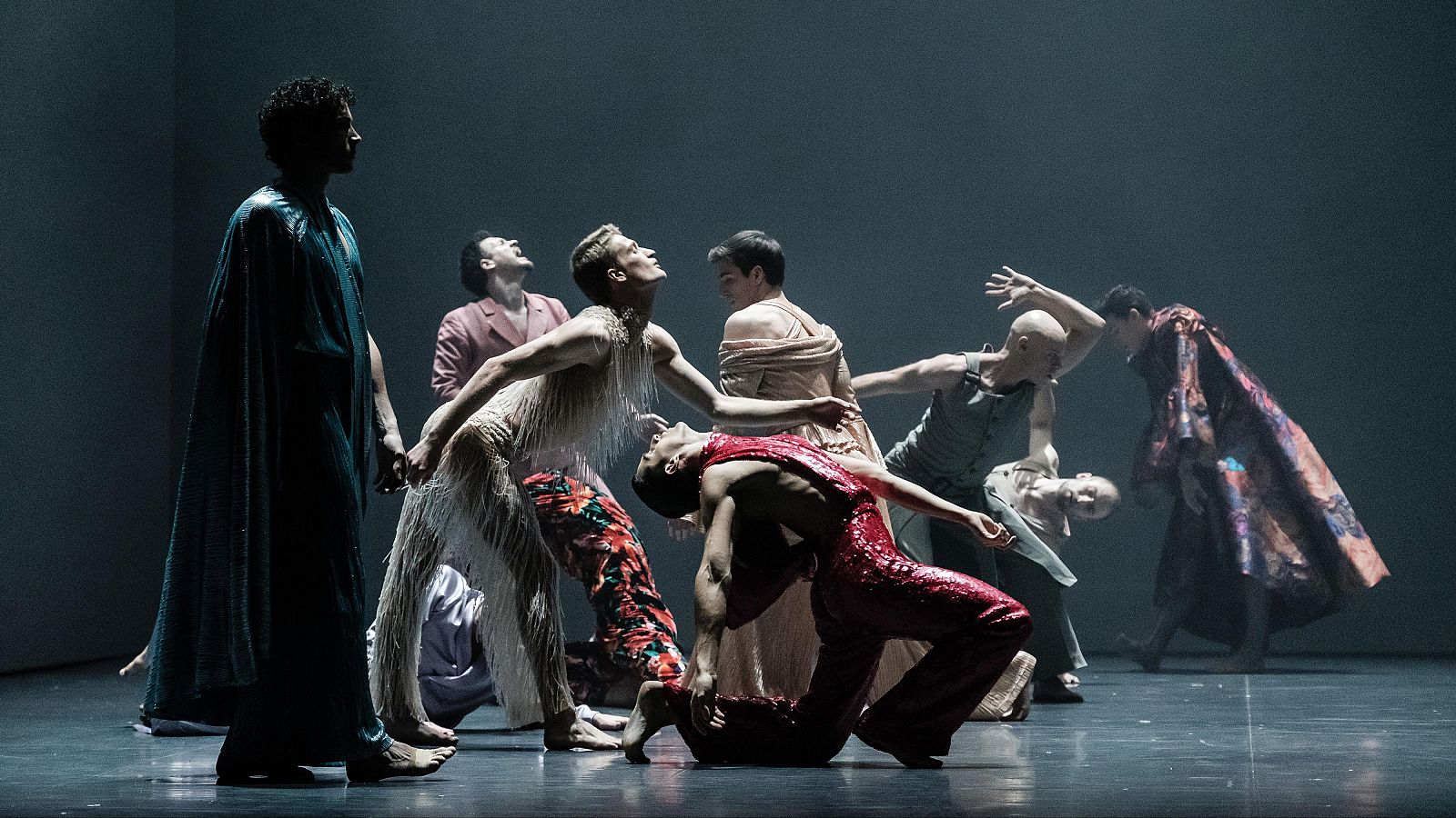 Ocho bailarines con vestuarios variados realizan una coreografía dinámica en un escenario oscuro e iluminado.  Poses y expresiones sugieren una exploración de las emociones humanas.