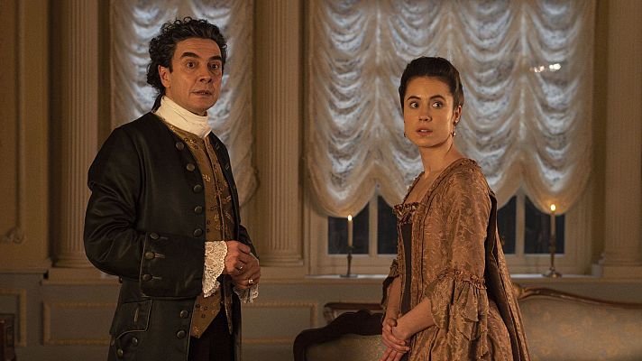 Dos personajes de época, él con chaqueta oscura y chaleco dorado, ella con vestido marrón dorado, conversan en una habitación elegante con candelabros encendidos.