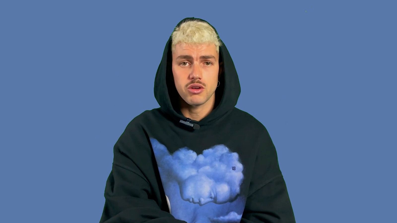 Cantante con sudadera negra y capucha sobre fondo azul.  Expresión neutra, cabello rubio platino corto y fino bigote.