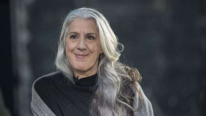 Plano general de Aitana Sánchez-Gijón con cabello gris suelto, vestida con ropa oscura y una prenda gris clara, mostrando una expresión serena.