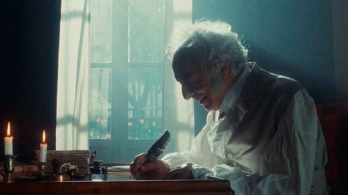 Hombre de edad avanzada escribiendo con pluma en escritorio antiguo, rodeado de velas y libros.  Imagen de documental.