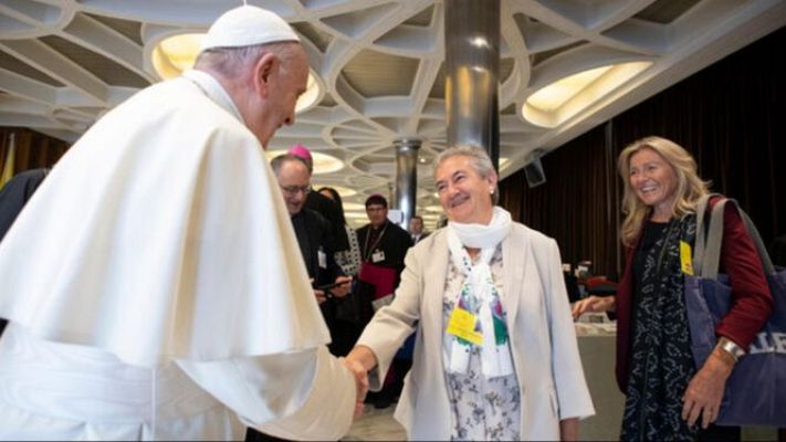 El Papa Francisco saluda a una mujer, quien viste un conjunto claro y bufanda, en una sala con columnas.  Se ven otras personas en el fondo.