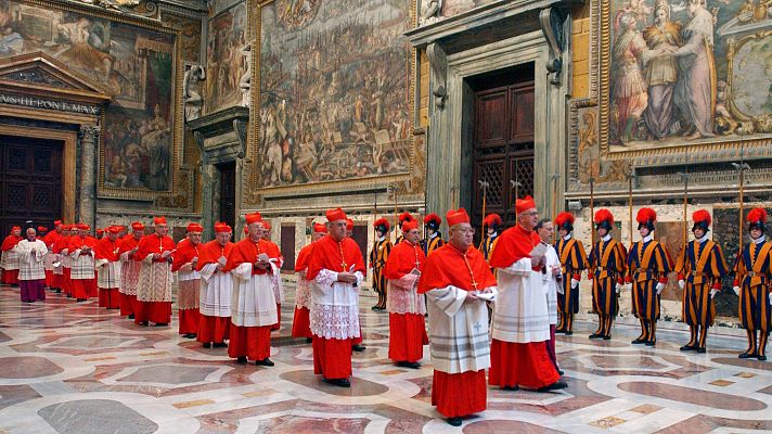 Procesión de cardenales en túnica roja y birrete en un salón del Vaticano con frescos y suelo de mármol.  Presentes también guardias suizos y clero en túnicas blancas.