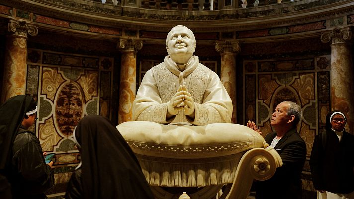 Estatua de papa orante rodeada de personas rezando, posiblemente en una iglesia. Ambiente de oración y expectación.