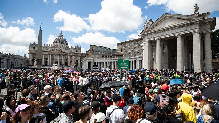 Multitud en la Plaza de San Pedro, con la Basílica al fondo y un letrero verde señalando una dirección.  Se ven paraguas y vallas.