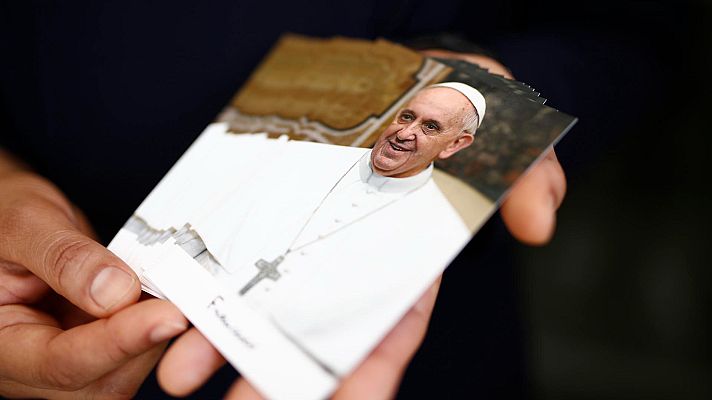 Manos sujetan fotos del Papa Francisco en atuendo blanco y cruz pectoral. Fondo interior claro con detalles dorados.  Imagen relacionada con su legado en El Salvador.