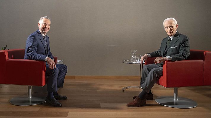 Entrevista formal entre dos hombres en sillones rojos. Uno viste traje azul marino y el otro traje gris.  Ambiente tranquilo.