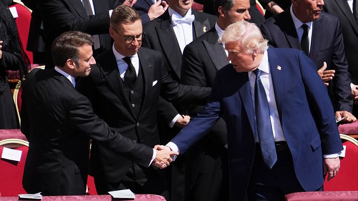 Macron y Trump, en trajes oscuros, se dan la mano en un funeral.  Asistentes con vestimenta formal al fondo.
