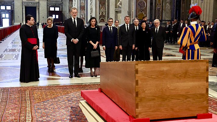Reyes de España en funeral de Estado.  Presentes ante féretro en espacio amplio con suelo de mármol y alfombra.  Acompañados de otras personas vestidas de negro.