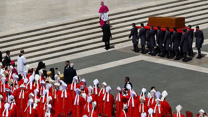 Tras la misa fúnebre, el féretro de madera es retirado de la Plaza de San Pedro por personal en uniforme. Cardenales en sotanas rojas observan, algunos usando sus móviles.