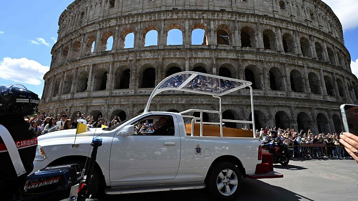 Cortejo fúnebre con ataúd visible en vehículo adaptado, escoltado por la policía, pasa frente al Coliseo romano entre aplausos de la multitud.