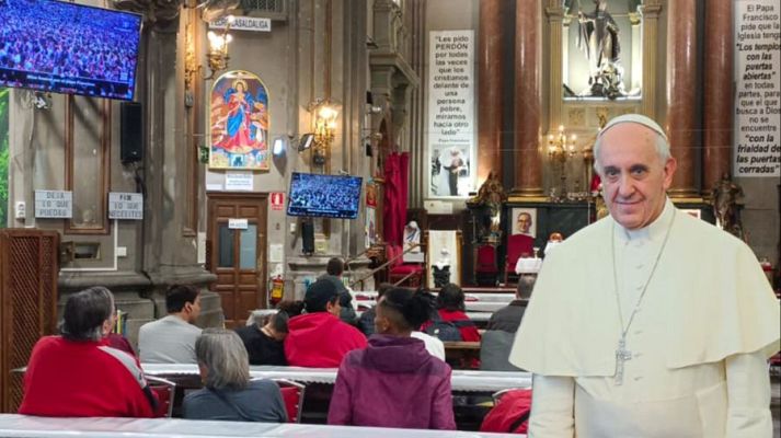 Imagen del Papa Francisco en una iglesia, con letreros y pantallas mostrando una multitud.  Viste de blanco y mira a la cámara.