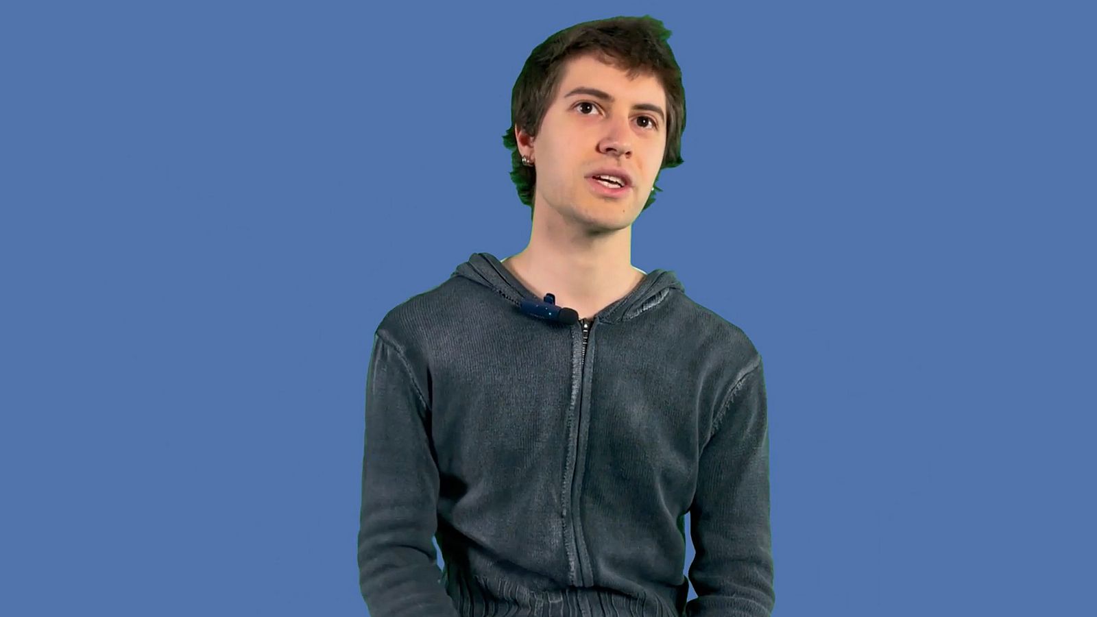 Joven con micrófono de solapa hablando en un programa de grabación. Fondo azul liso.