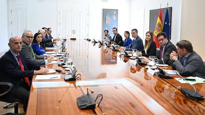 Reunión en sala oficial con doce personas alrededor de una mesa, micrófonos y teléfonos presentes.  Ambiente serio.