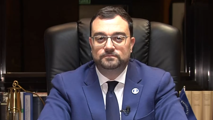 Hombre en traje azul, camisa blanca y corbata, sentado en silla de oficina, posiblemente presentando un informativo.  Pin en la solapa, estantería con libros al fondo.