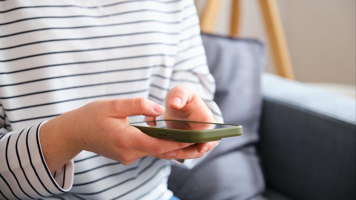 Persona con camiseta a rayas usando un teléfono móvil con funda verde oliva. Fondo con sofá gris y madera.  Situación de apagón.