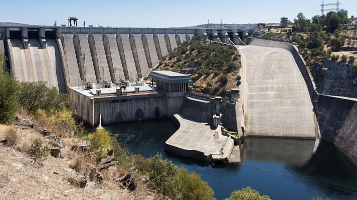Presa de hormigón con compuertas, central hidroeléctrica y torres de alta tensión visibles. Flujo de agua controlado.  Almacenamiento de energía.