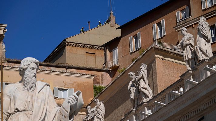 Trabajadores con cascos en un tejado Vaticano realizan tareas de mantenimiento o preparación, con estatuas en primer plano y un cielo despejado.