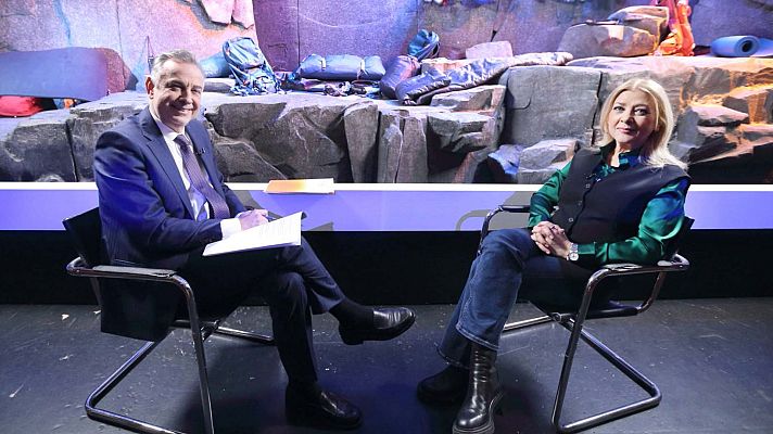 Entrevista en plató con Vicente Vallés y una mujer con camisa verde y chaleco. Decorado rocoso con material de escalada. Iluminación tenue, azulada y violeta.