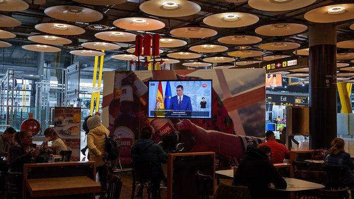 Personas en sala de espera viendo discurso televisado del presidente, acompañado de intérprete de lengua de signos.  Ambiente de espera, posiblemente relacionado con un anuncio importante.