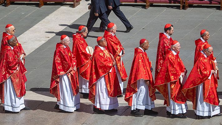 Cardenales en sotanas blancas y capas rojas doradas desfilan por una plaza de piedra en una procesión.