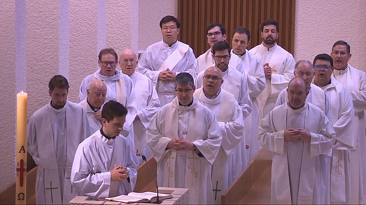 Grupo de clérigos en interior claro con paneles de madera, uno leyendo un libro y una vela con alfa y omega visible.  Posible momento de oración o estudio.