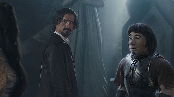 Karra Elejalde, con larga cabellera oscura y chaqueta negra, junto a Hei Buh, personaje animado con armadura y cadena, en un ambiente oscuro y misterioso.