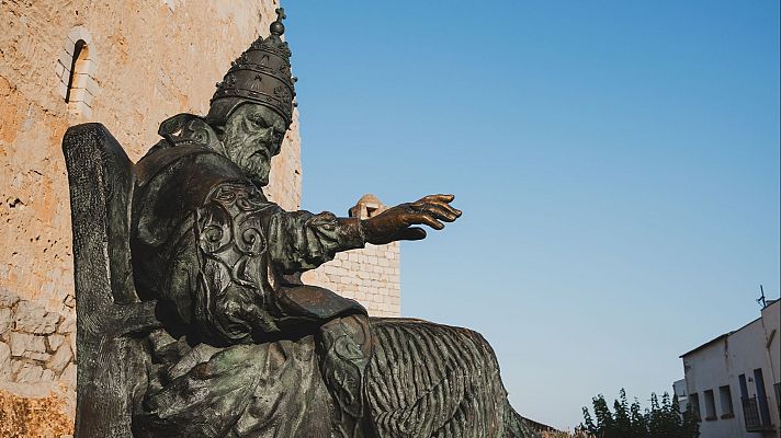 Estatua de bronce de figura religiosa con mitra y túnica, sentada en un trono, en un espacio abierto con edificios de piedra al fondo.
