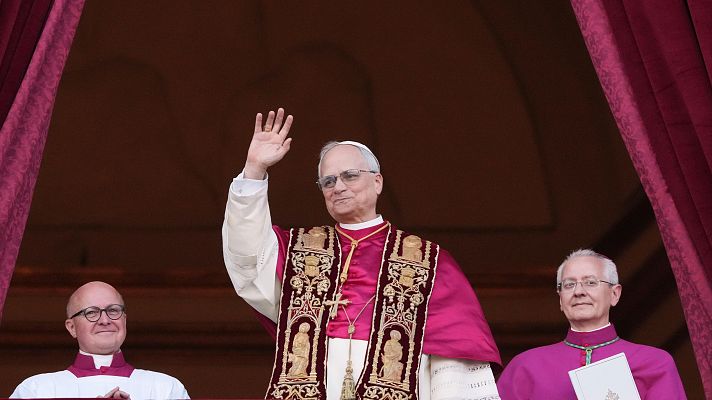 El Papa Francisco, con vestimenta papal y gafas, saluda desde un balcón. Dos hombres con atuendos religiosos le acompañan.