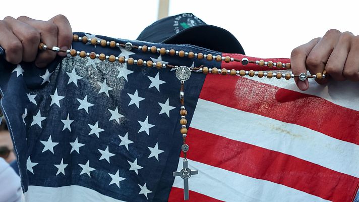 Persona con gorra sostiene bandera de EEUU y rosario; rostro parcialmente oculto.  Posible manifestación de fe o patriotismo.