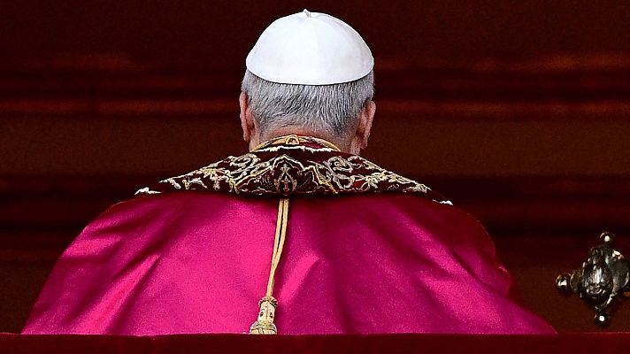 Hombre de espaldas, vestido con capa magenta, cuello dorado y solideo blanco.  Atuendo religioso que sugiere un alto cargo eclesiástico.