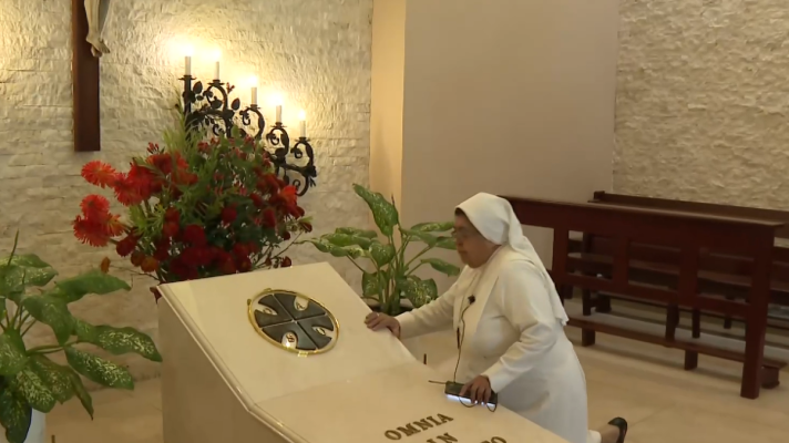 Mujer con micrófono junto a un atril con texto en latín en una capilla.  Se observa un arreglo floral cercano.