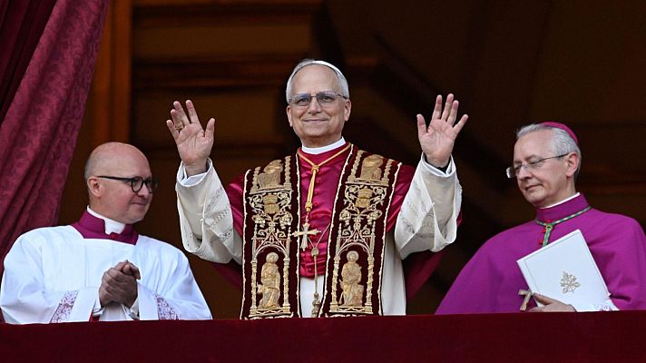 El Papa León XIV, en casulla roja y dorada, saluda desde un balcón; clérigos lo acompañan, uno aplaudiendo y otro con un documento.
