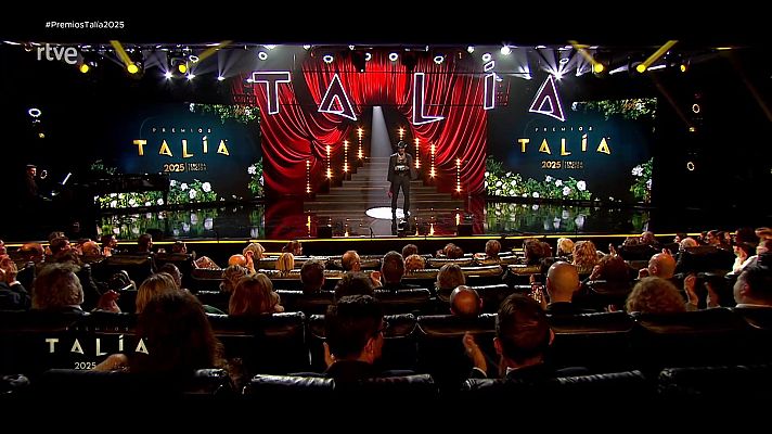 Gala de los Premios Talía 2025 retransmitida por La 2.  Presentador en escenario con cortinas rojas y pantallas mostrando el logo del evento y flores. Público aplaudiendo.