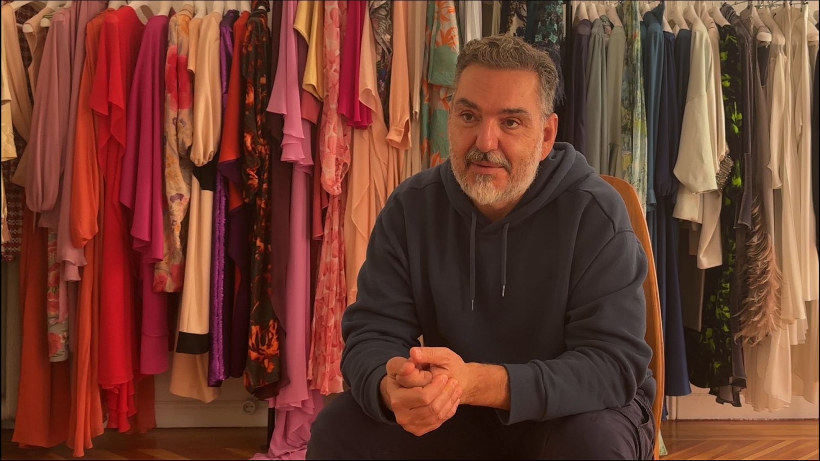 Diseñador sentado con sudadera azul, ante un perchero con ropa vibrante.  Posible escena de entrevista o programa.