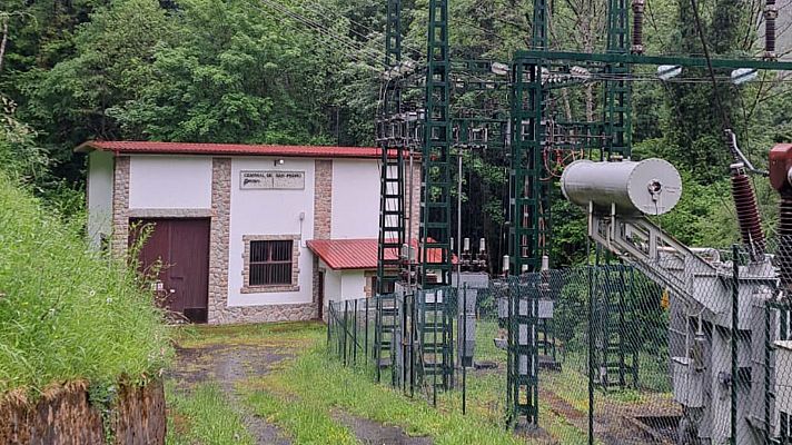 Pequeña construcción blanca con techo rojo, puerta marrón y ventana enrejada, ubicada en entorno rural con abundante vegetación e instalación eléctrica cercana.
