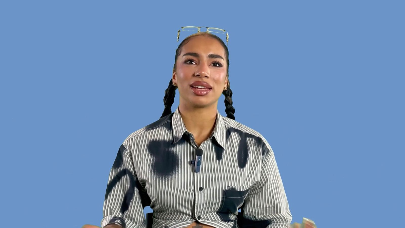 Cantante con camisa a rayas, gafas y dos trenzas, usando un micrófono lavalier en una entrevista o grabación sobre fondo azul.