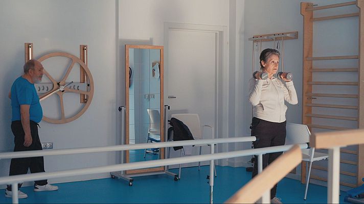 Dos personas mayores realizan ejercicios de rehabilitación: un hombre en una rueda de pared y una mujer con pesas. La sala incluye barras paralelas y espaldera.