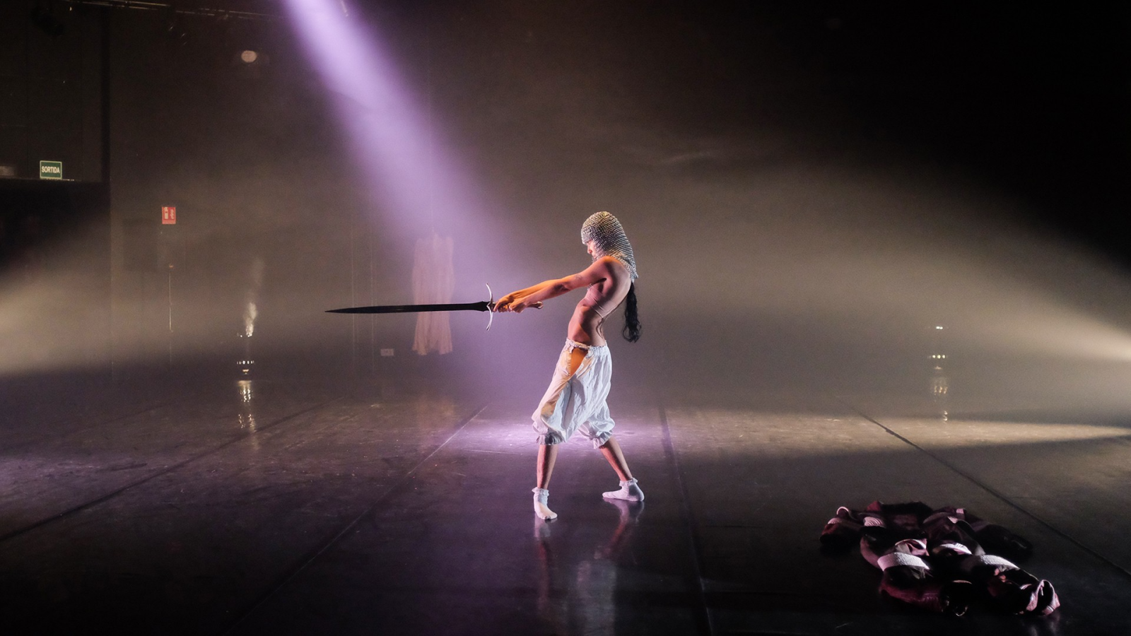 Artista en escena con cota de malla y espada, iluminación dramática.  Prenda oscura en el suelo.