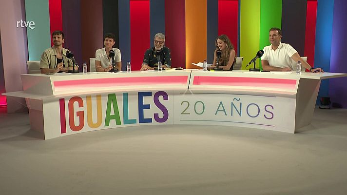 Plató de televisión con seis personas alrededor de una mesa blanca con el logo \"IGUALES 20 AÑOS\". Micrófonos individuales y botellas de agua sobre la mesa. Fondo con franjas verticales de colores.